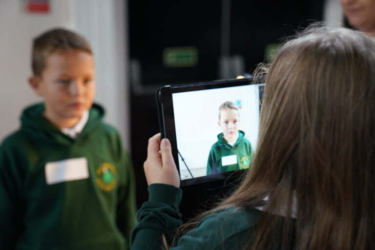 A young girl capturing a photo of a boy on a tablet device. 