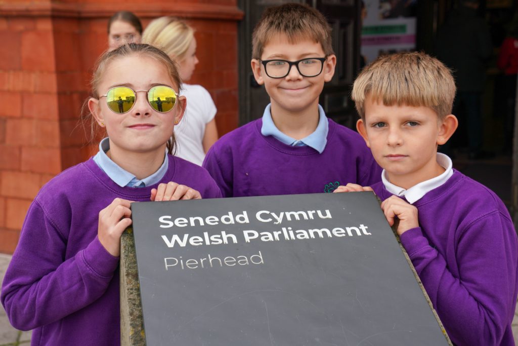 Tri plentyn yn gafael arwydd yn dweud "Senedd Cymru Welsh Parliament" o'u blaen