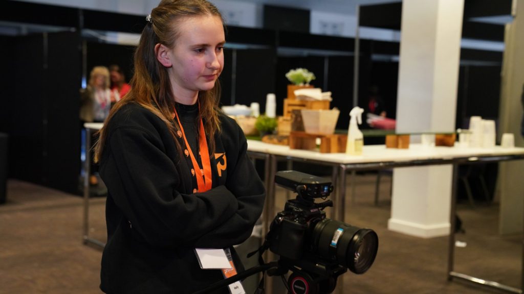 Molly, a young female employee, standing with her arms folded cross behind a professional camera on a tripod. 