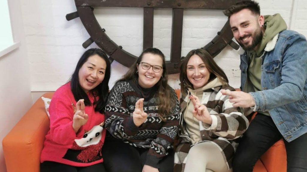 Four staff members sat on the sofa, smiling at the camera. They are holidng up one hand each with crossed fingers, to imitate the National Lottery Community Fund logo. 