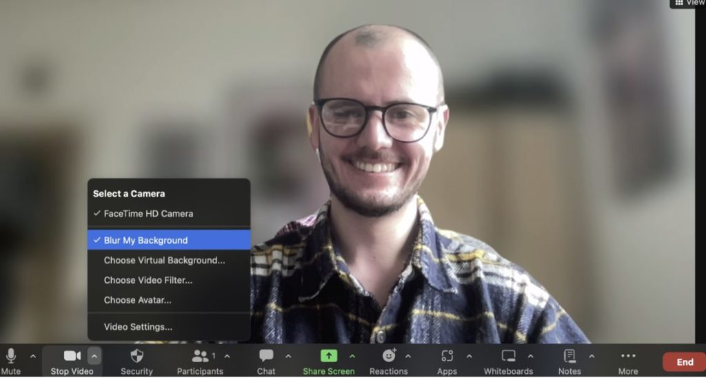 Screenshot of a man smiling on Zoom, hovering over the 'Blur my background' button on the toolbar. 