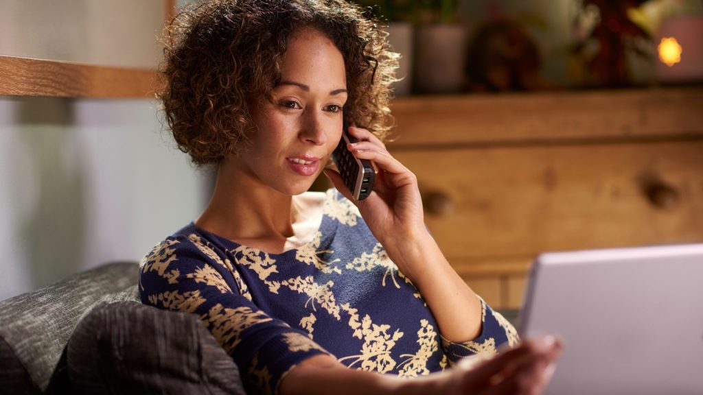 Woman on sofa on movile phone, with laptop open in front of her. Her face shows a thinking expression.