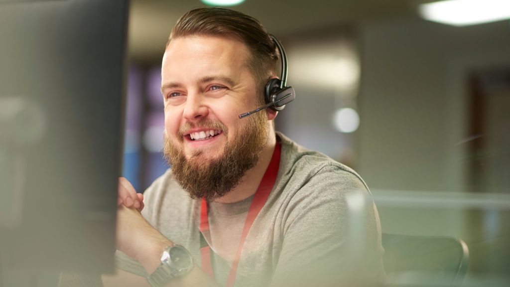 Adult male wearing a headset in an office environment. He is smiling into the distance whilst on the call.