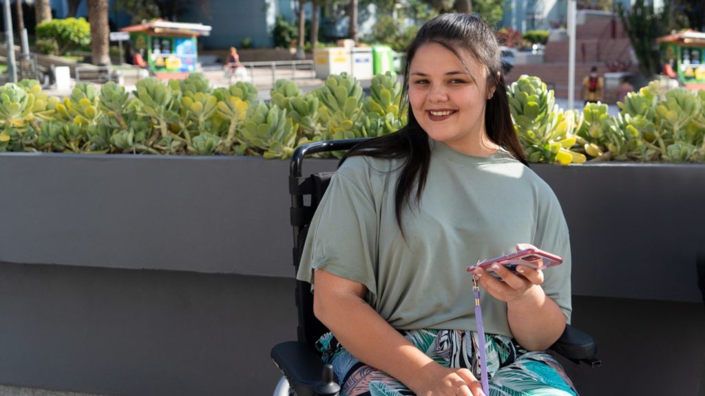 Young adult female sat in a wheelchair outdoors. In her hand is a mobile phone.