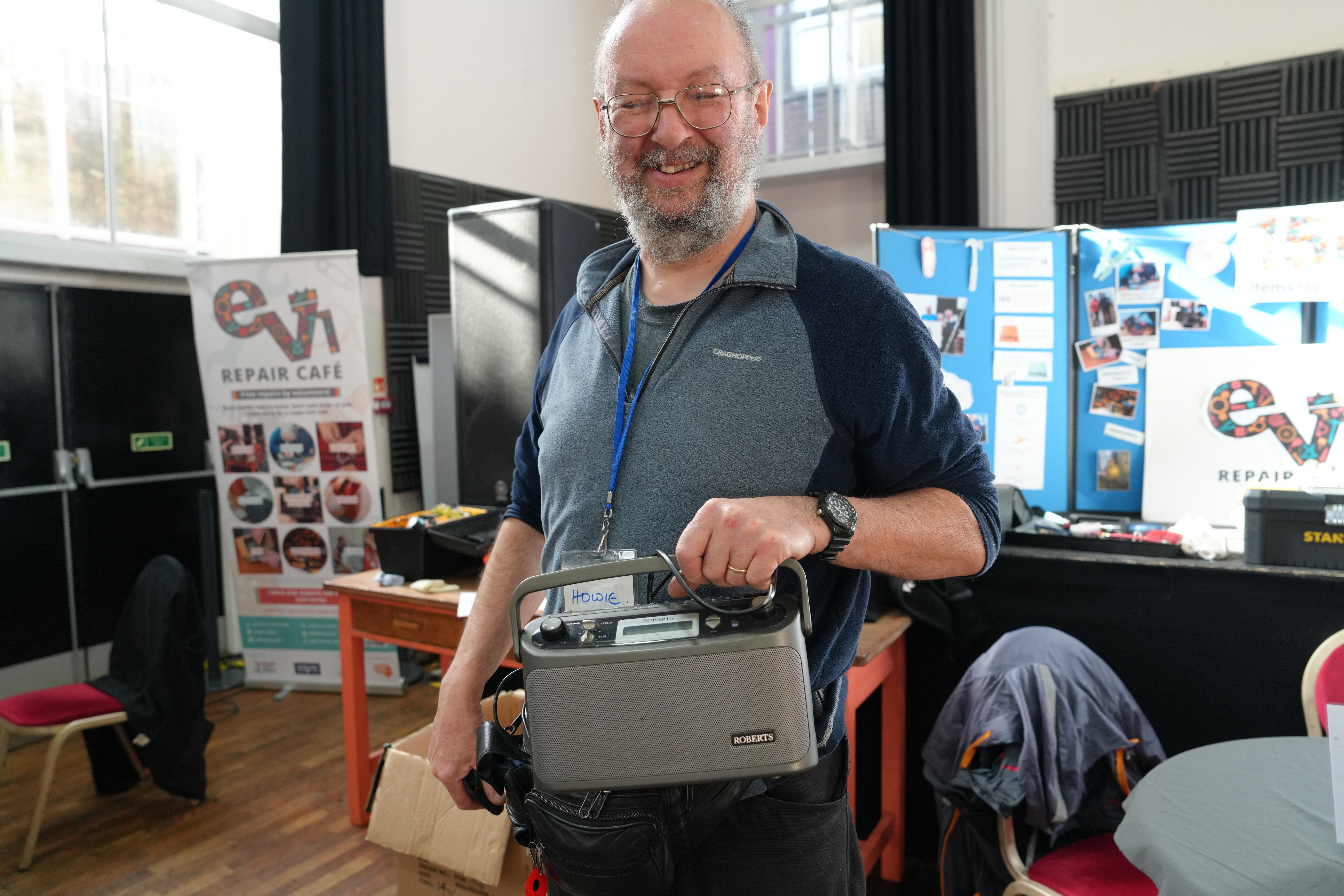 A man smiling and holding a radio, standing in a room with "Repair Café" banners and posters in the background.