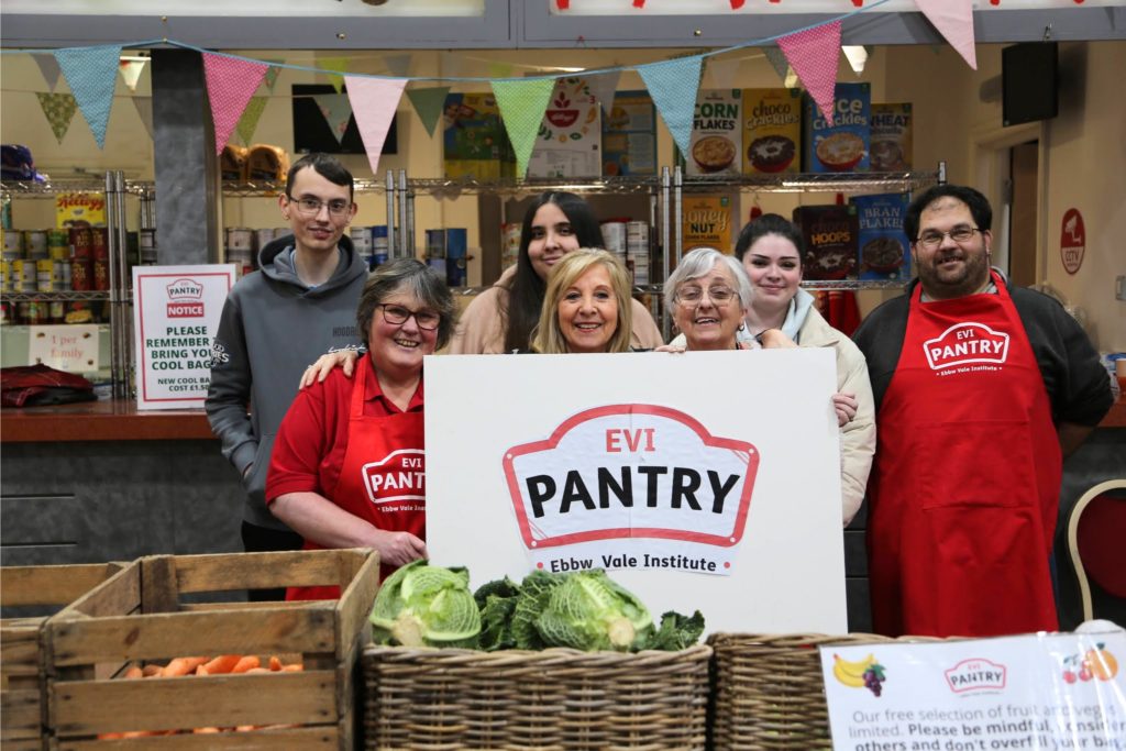 A group of six people smiling and holding a sign for EVI Pantry inside a food pantry, with shelves of groceries and crates of vegetables in the background.