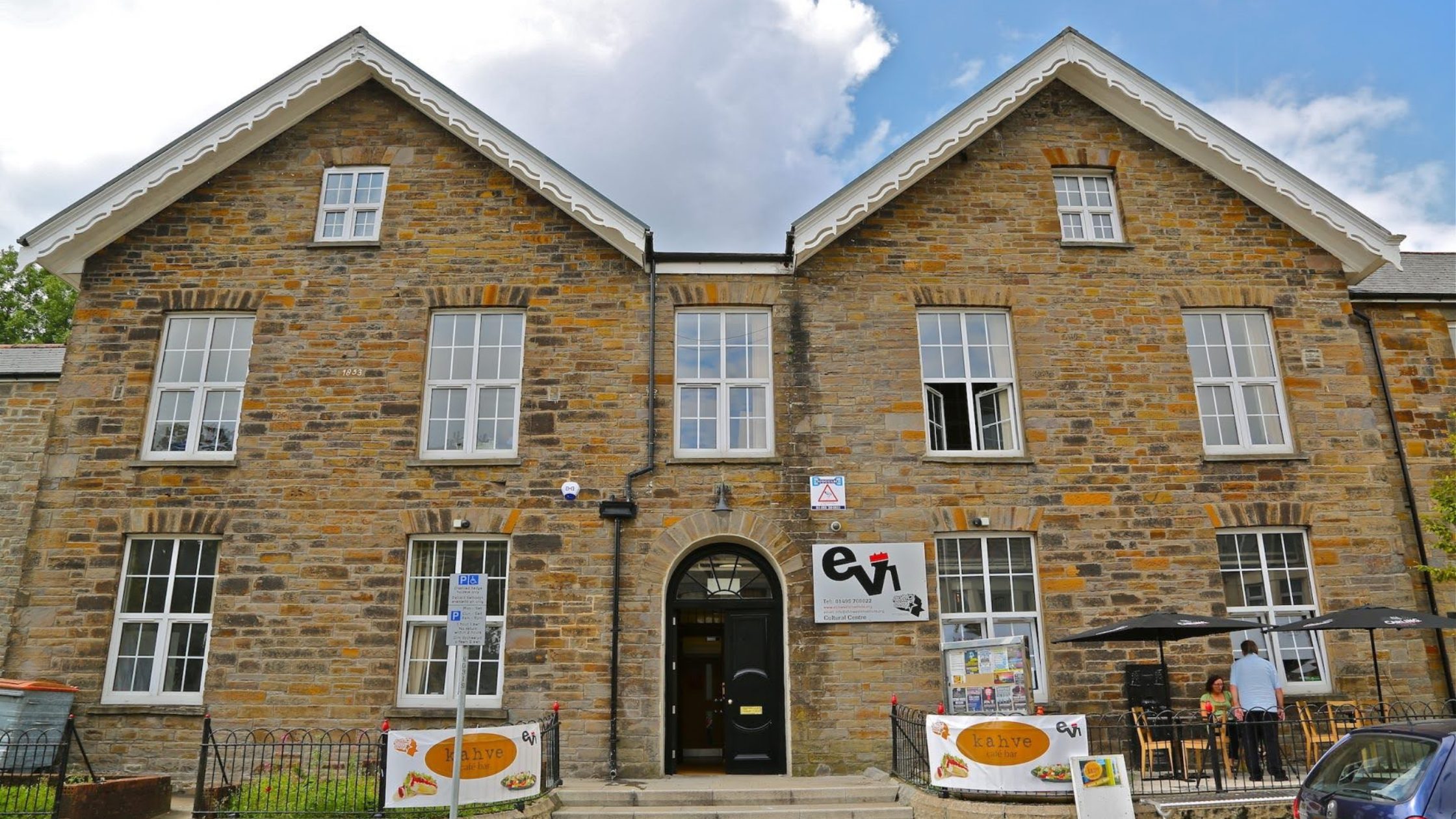 A historic stone building with two peaked roofs, called the Ebbw Vale Institute. Signs and outdoor seating are visible near the entrance, along with banners advertising offerings for the community.