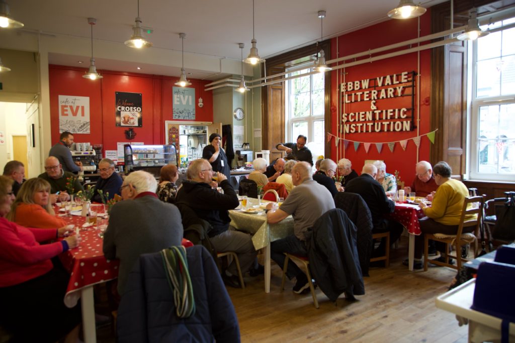 Large group of people are seated and conversing at tables in a brightly lit room decorated with posters and bunting.