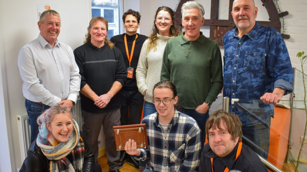 Group of ProMo Cymru staff stood together, smiling for a photo with a wooden plaque to showcase the Bronze Youth Work Award.