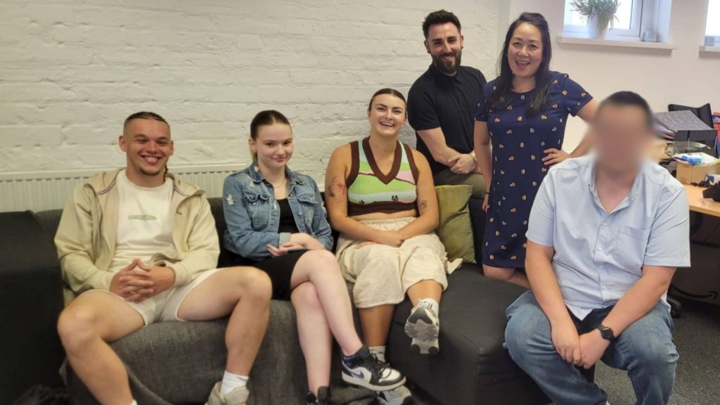 Group of 6 young people and staff members smiling on a grey sofa together