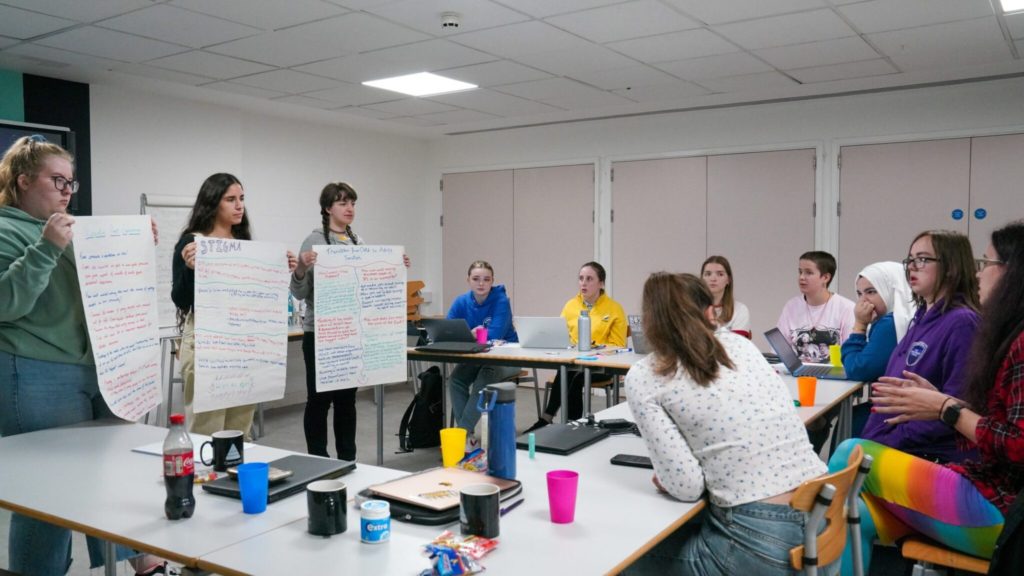 Three staff members stood presenting on flip chart paper to a room of young people. 