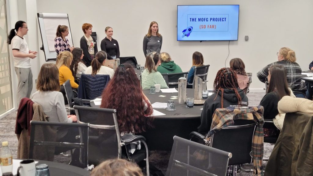 Group of young people stood at the front of a conference room, presenting to professionals sat around circular tables