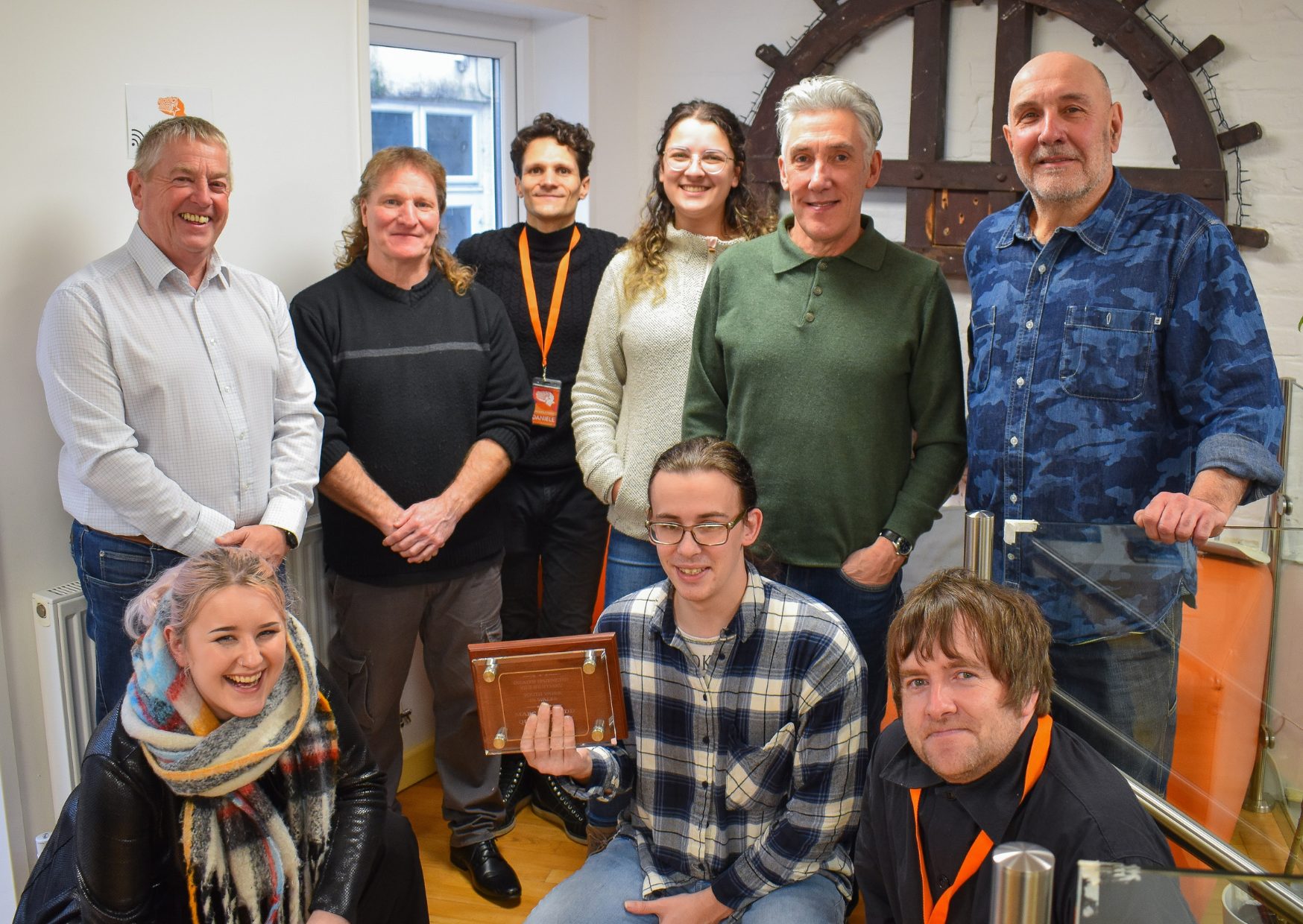 Group of ProMo Cymru staff stood together, smiling for a photo with a wooden plaque to showcase the Bronze Youth Work Award.