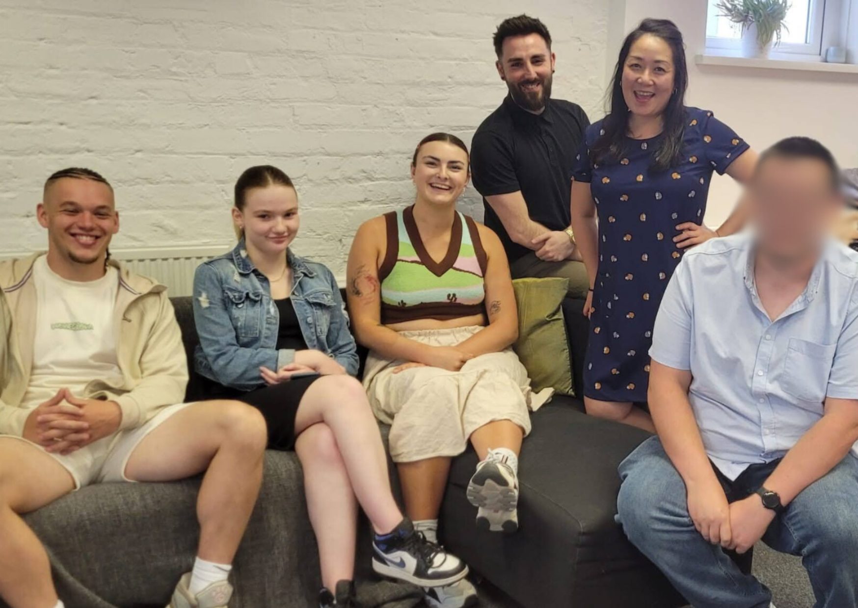 Group of 6 young people and staff members smiling on a grey sofa together