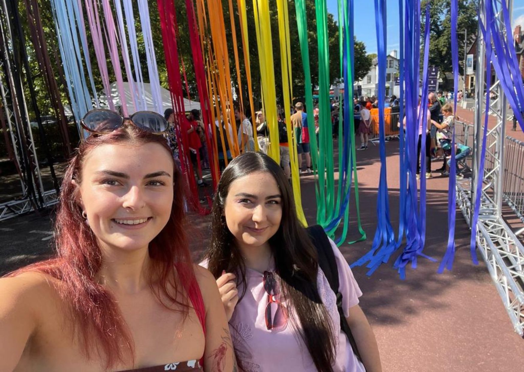 Two women talking a selfie in front of a rainbow background made of ribbons