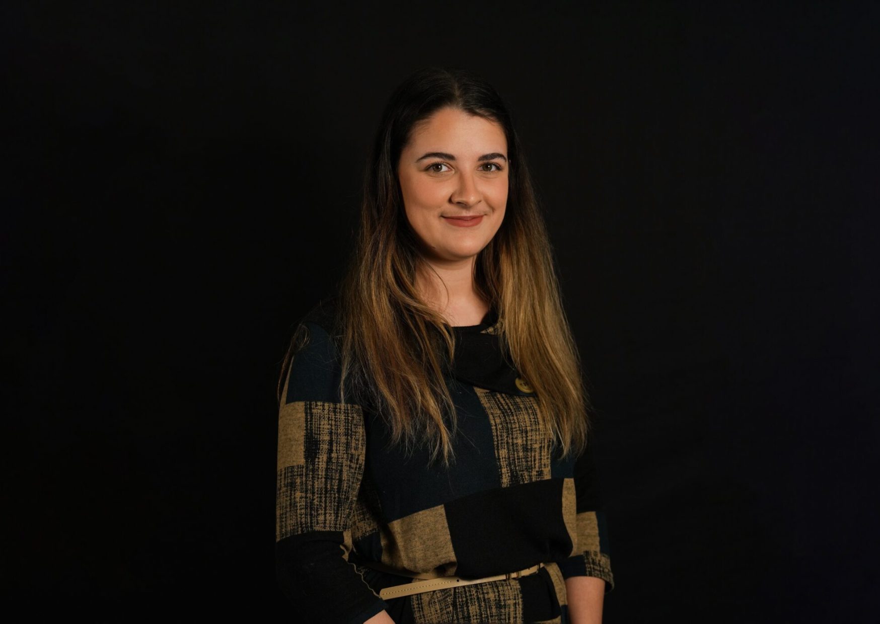 Portrait of Simran, a young female employee with long light brown hair with highlighted tips. She wears a black and brown chequered dress.