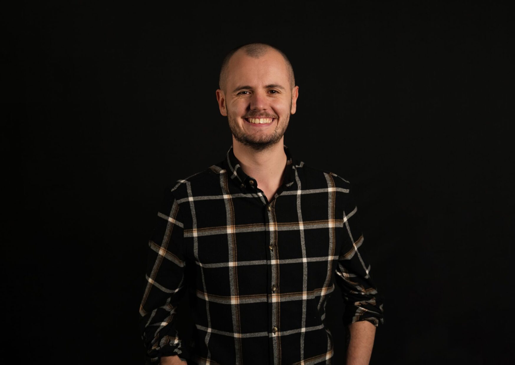 Portrait of Joe, wearing a black long sleeved shirt with white checks, rolled up to his elbows. He is smiling at the camera.