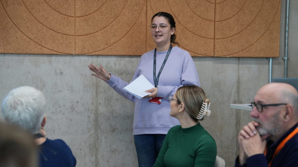 Image of Simran wearing a light purple sweater. She is presenting in front of a room of engaged staff. 