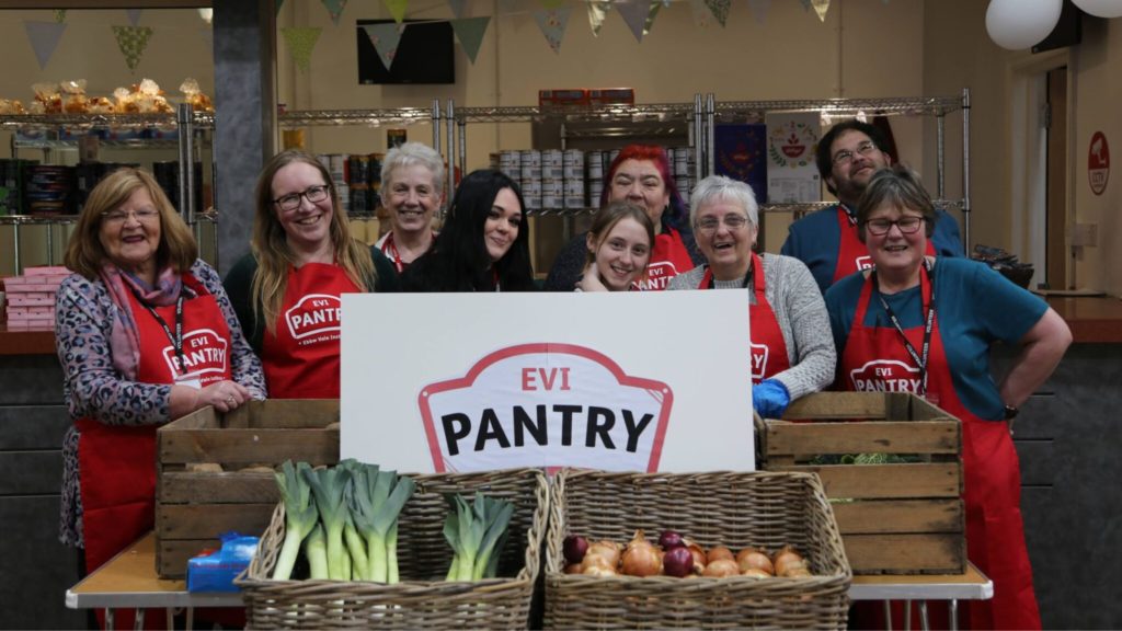 Image of 9 staff and volunteers stood behind a sign that reads EVI pantry. In the foreground, there are boxes full of fresh foods such as leeks and onions.