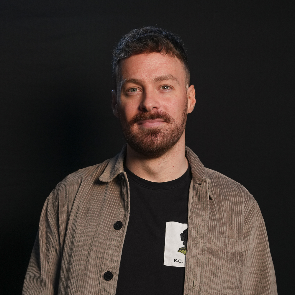 Portrait of Adam, a brown haired male with a moustache and a short beard. He is wearing a black t-shirt and brown shirt, and is smiling at the camera.