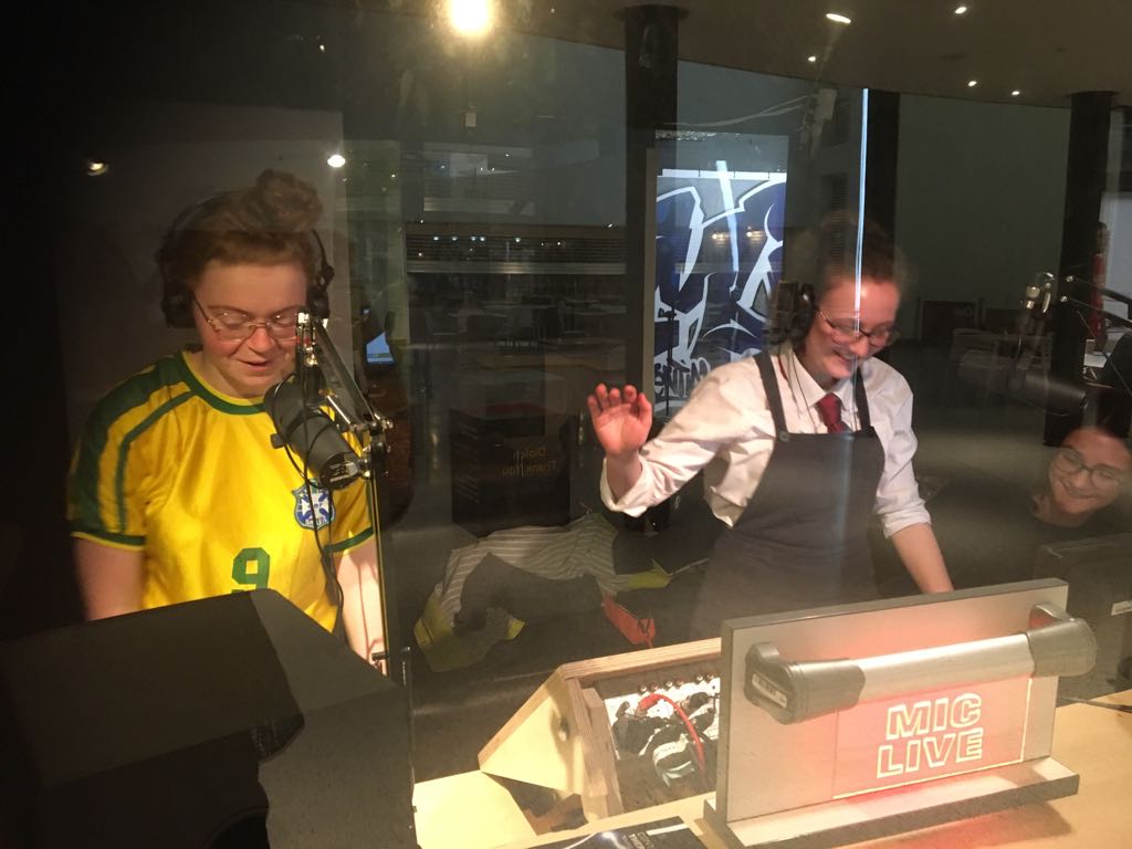 Two girls behind the glass of a recording studio talking into microphones