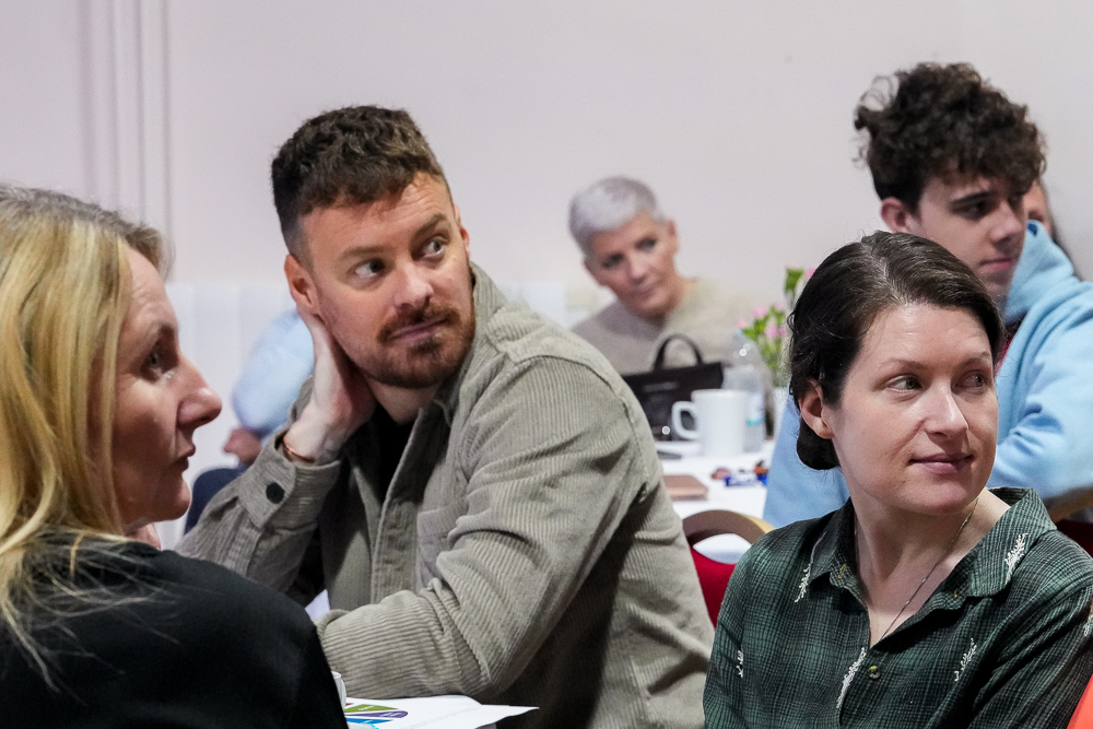 People in a room all looking in one direction (at a speaker out of shot) The main figure at the centre of the image is a male with a short trimed beard in a corduroy green shirt with his palm resting on the back of his neck.