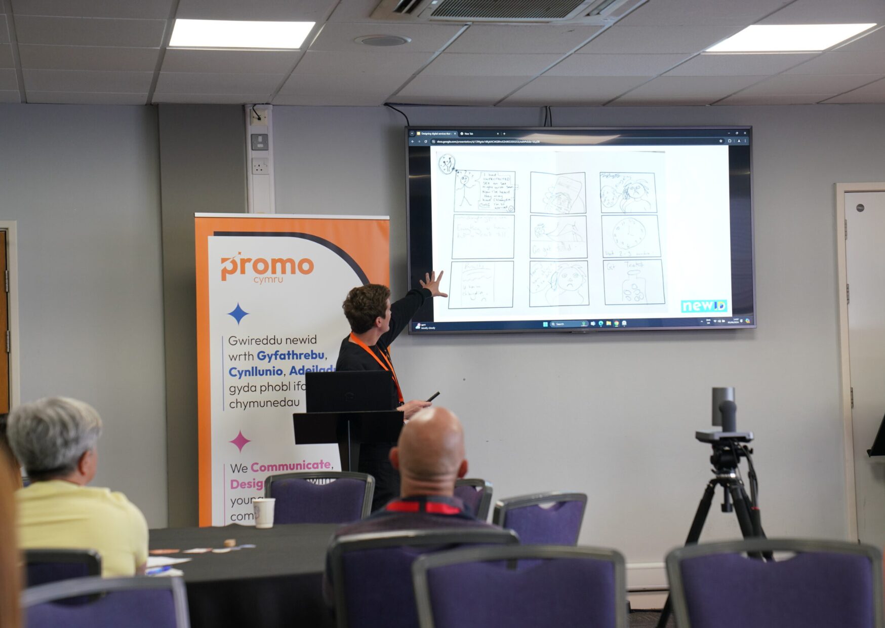 Person pointing to a screen teaching a room of people. There is a ProMo Cymru pull up banner behind him.