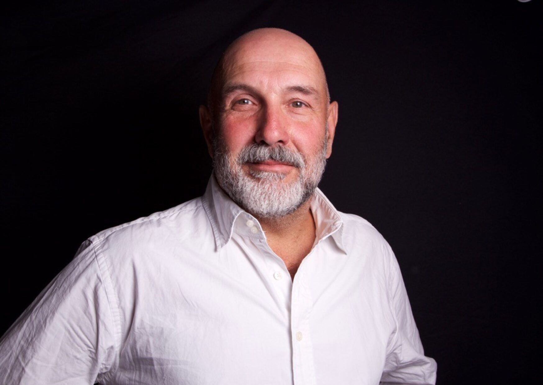 Picture of the top half of a bearded man in a white shirt smiling at the camera. The background is black. This is the Chief Executive of ProMo Cymru, Marco Gil-Cervantes.