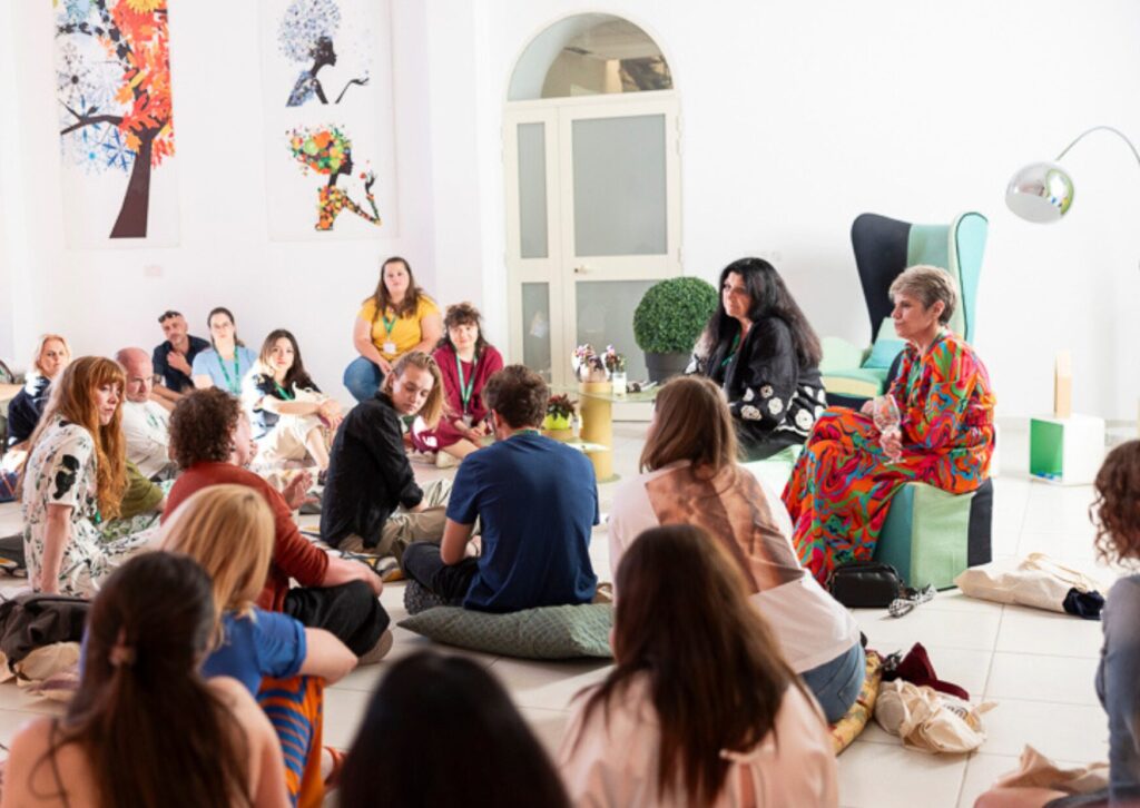 Attendees at the convention sit in a bright, white room with colorful art on the walls, participating in a relaxed group discussion.