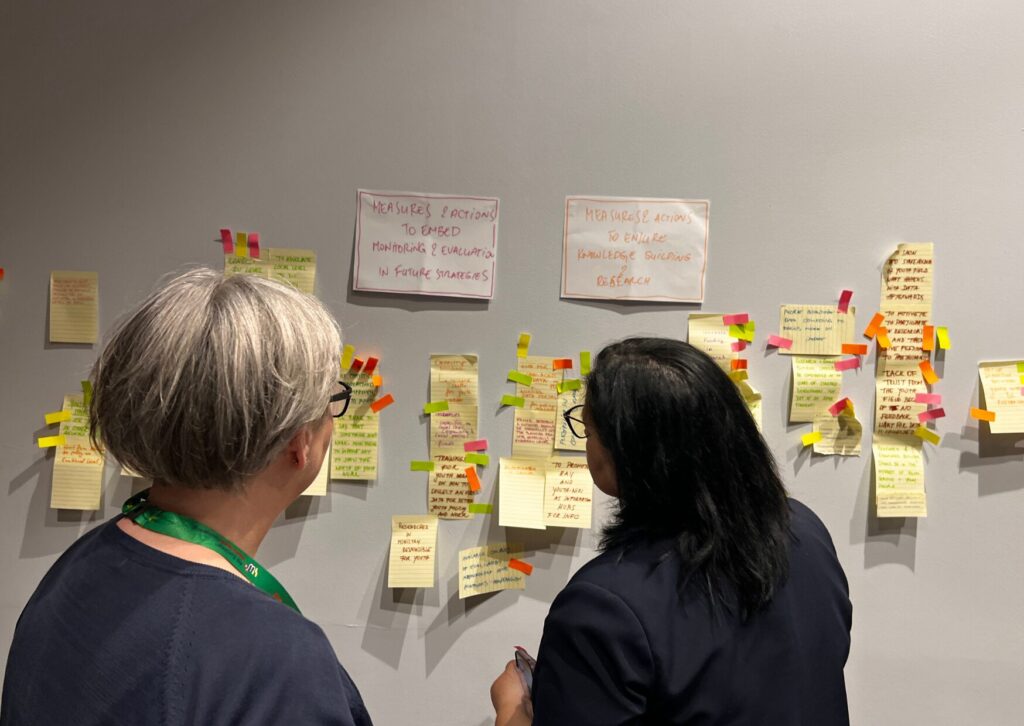 Two conference attendees stand in front of a wall covered in yellow, pink, and orange sticky notes, a common sight at a workshop or brainstorming session.