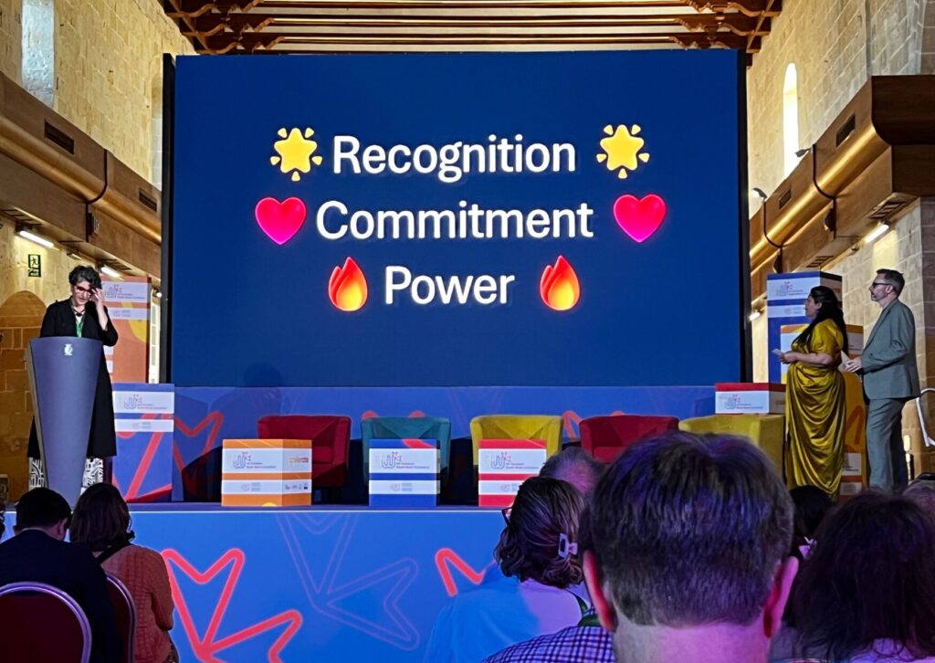 A speaker stands at a podium on a stage with a large screen behind them. The screen displays the words 'Recognition,' 'Commitment,' and 'Power,' each with a different emoji.