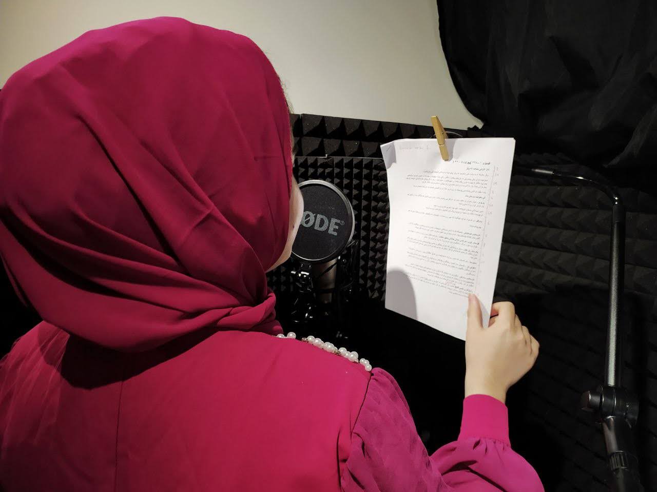 Close up image of the head and sholders of a young girl taken from behind. She is wearing a red top and hijab and is holding a piece of paper in her hand, talking into a rhode microphone