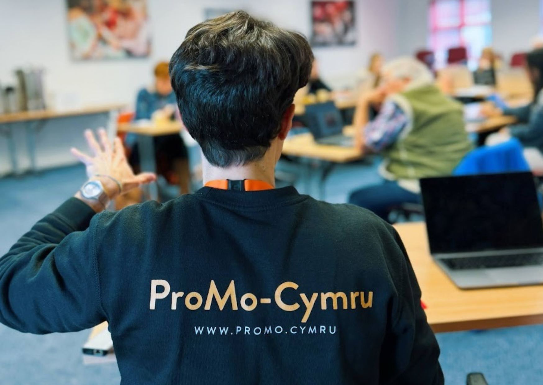 Photo of the back of a person in black jumper with the writing ProMo Cymru on it. They are presenting to a room of people sat on desks.
