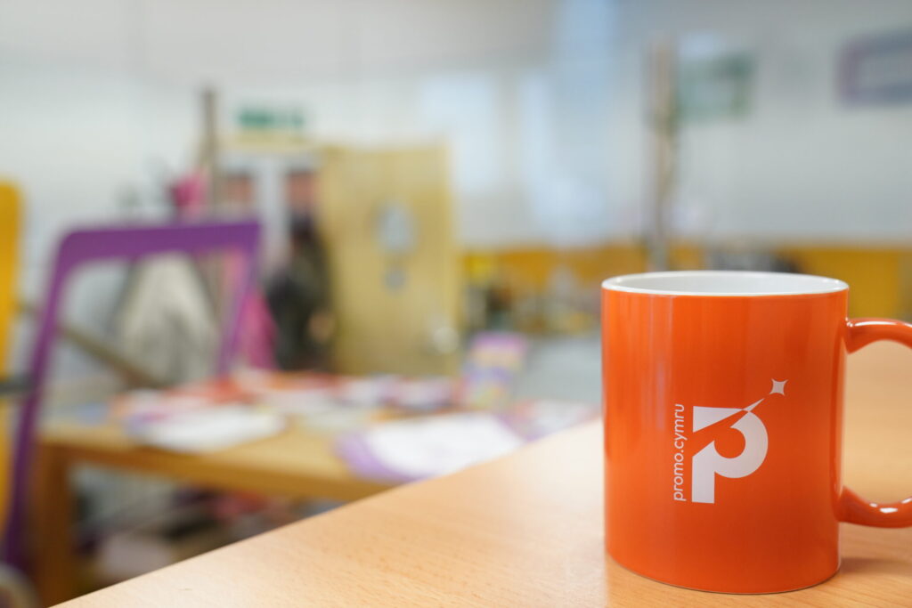 a photo of an orange mug on a desk. The background is blurred. The mug has a logo on it a P with a star and the words promo cymru small on the side of the p.