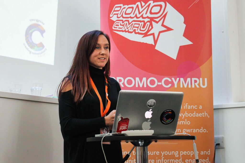 A young woman (Arielle) stood in front of a presentation screen with a ProMo Cymru pull out stand next to her and a laptop on the podium in front of her.
