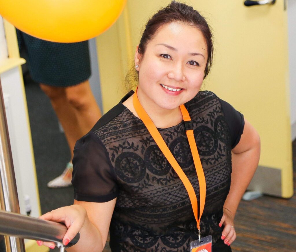 A young woman (Cindy) posing smiling at the camera wearing a black top and orange lanyard