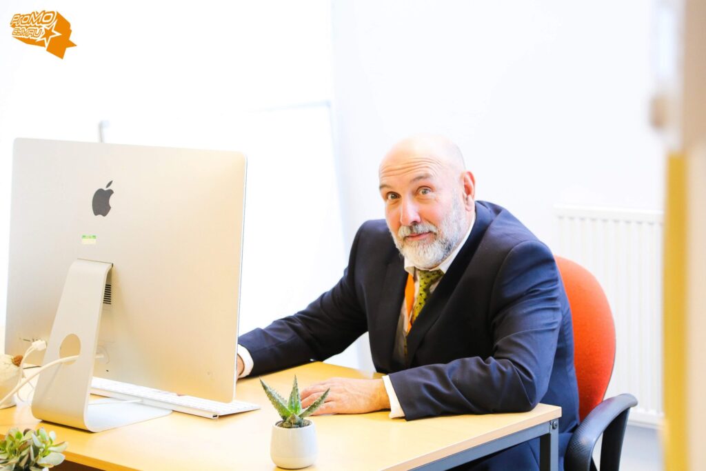 A bearded man in a suit (Marco) sat at a desk in front of a computer screen looking at the camera.
