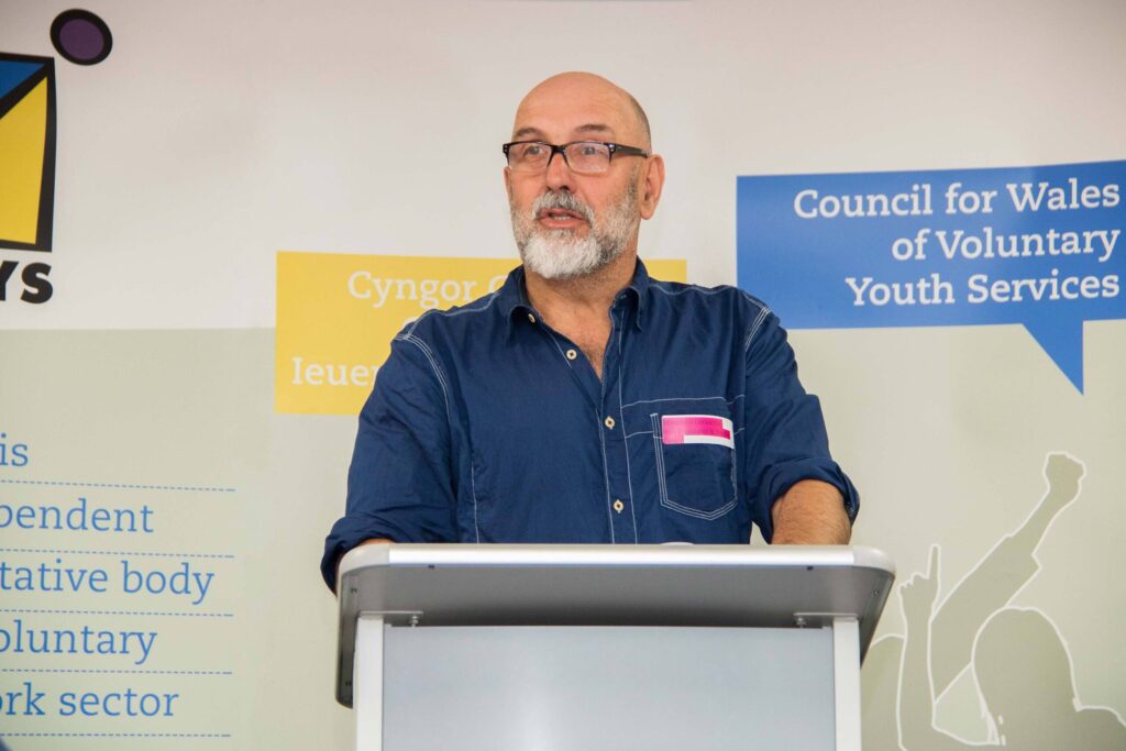 A bearded man (Marco) wearing a blue shirt and glasses stood in front of a podium