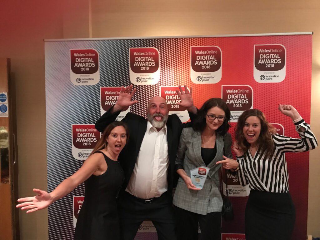 Four people (3 female and one male) stood in front of a Digital Awards background doing silly poses for the camera holding an award.