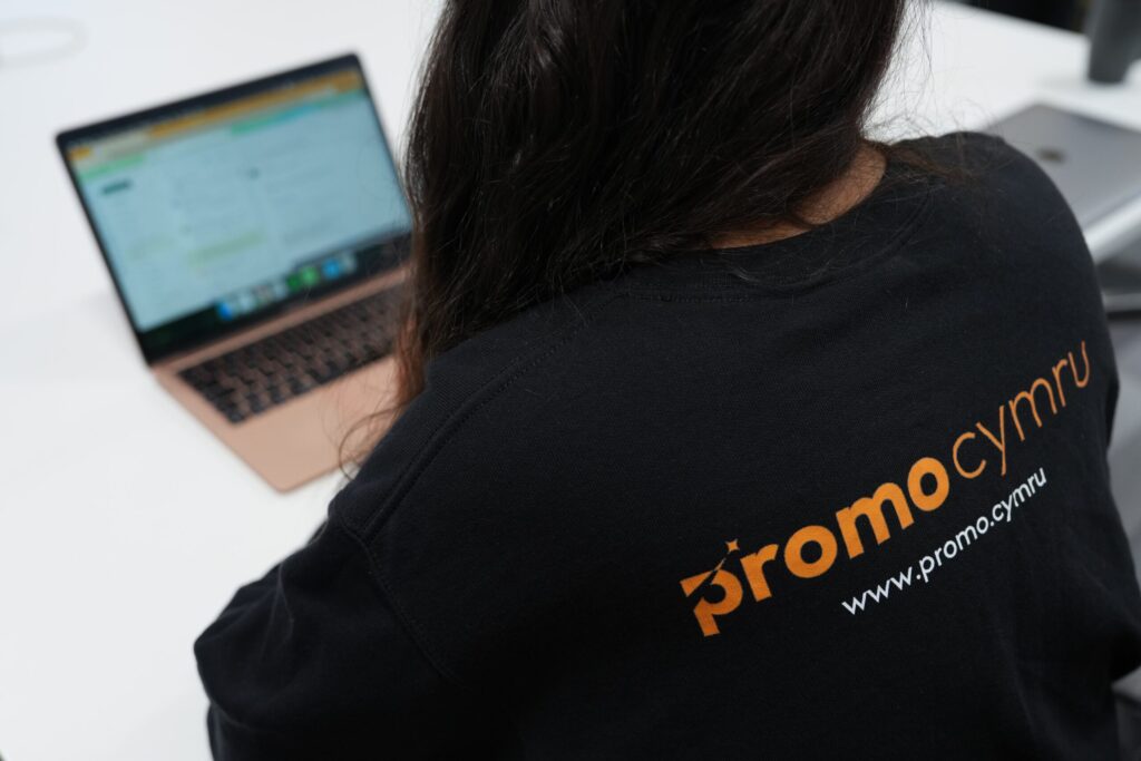 a photo from the back of someone on a laptop. They are wearing a black jumper with the words promo cymru on it - showing the new branding developed