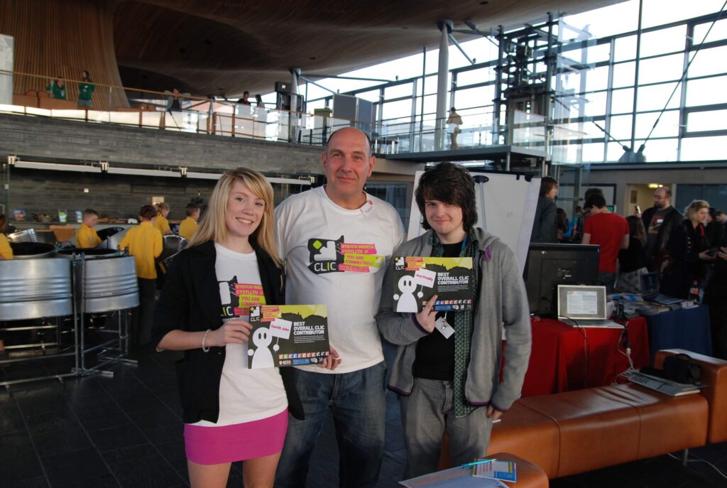 A man in a white tshirt (Marco) in the middle of two young people holding up a booklet at the Senedd building.