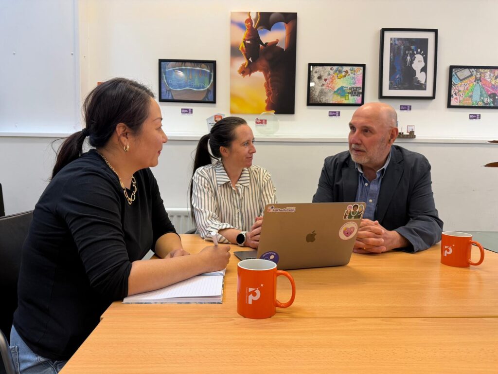 Three people (Cindy, Arielle and Marco) engaged in conversation sat at a desk with a laptop on it and two orange mugs with the letter P on them.