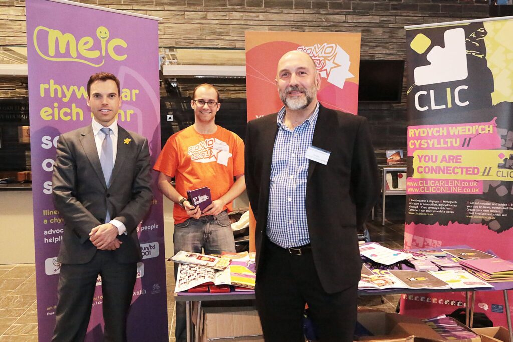 Three people stood on a stall with Meic, ProMo and CLIC banners behind them.
