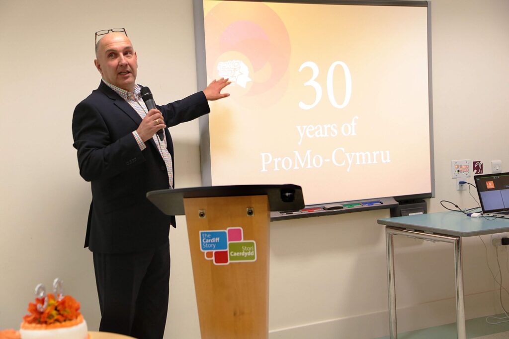 A man holding a microphone (Marco) in front of a podium pointing at a presentation on screen saying 30 years of ProMo Cymru