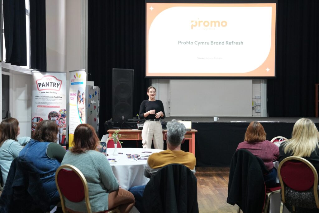 A person stoof in a big hall with a huge screen behind with the promo cymru logo and the words ProMo Cymru Brand Refresh on it. People are around tables listening to the presentation.