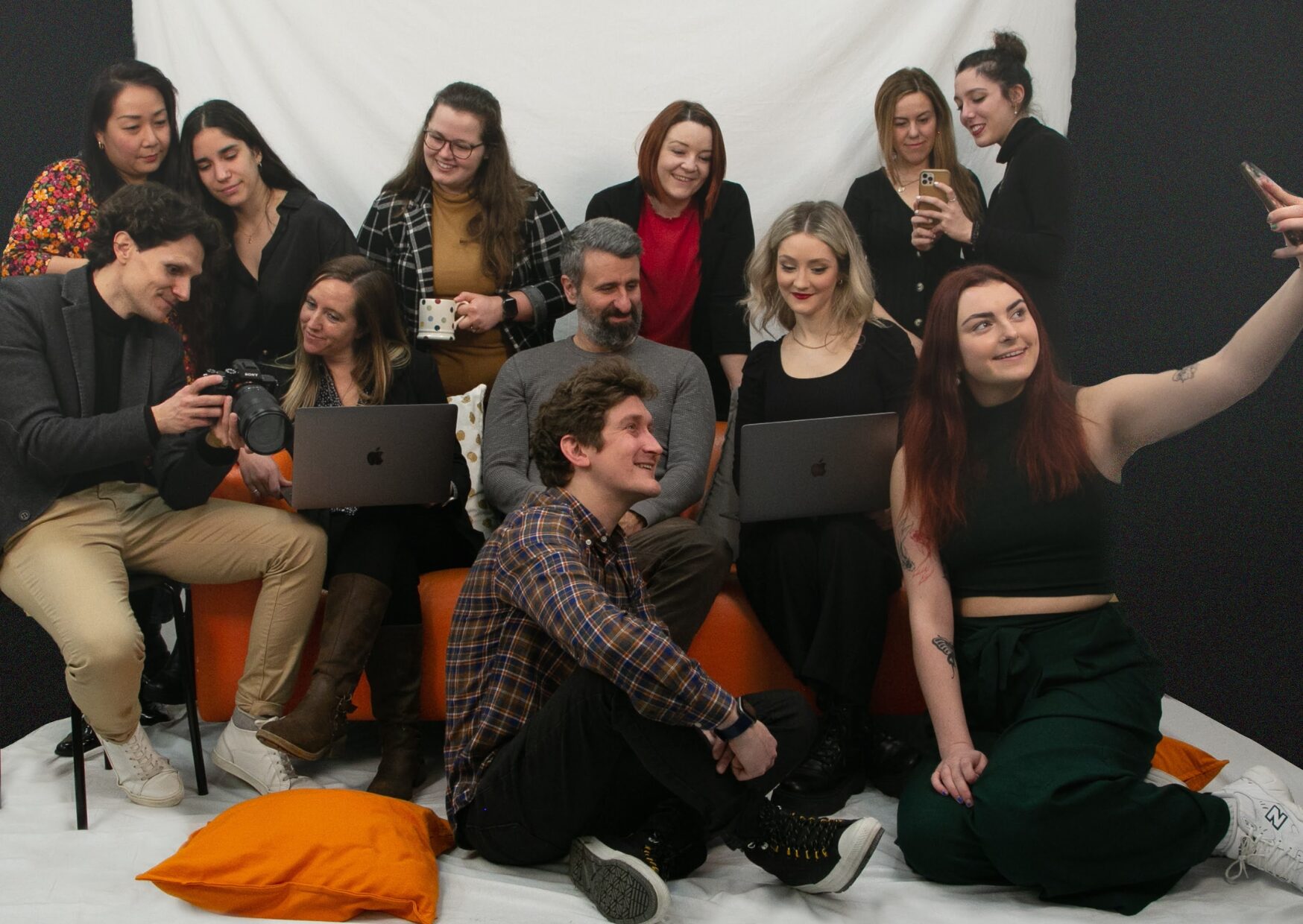 Group of people gathered on sofa, standing and sitting on floor. All looking at different devices together, smiling.