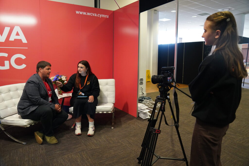 Female interviewing a male, sat on a white sofa. Person in foreground is filming them.