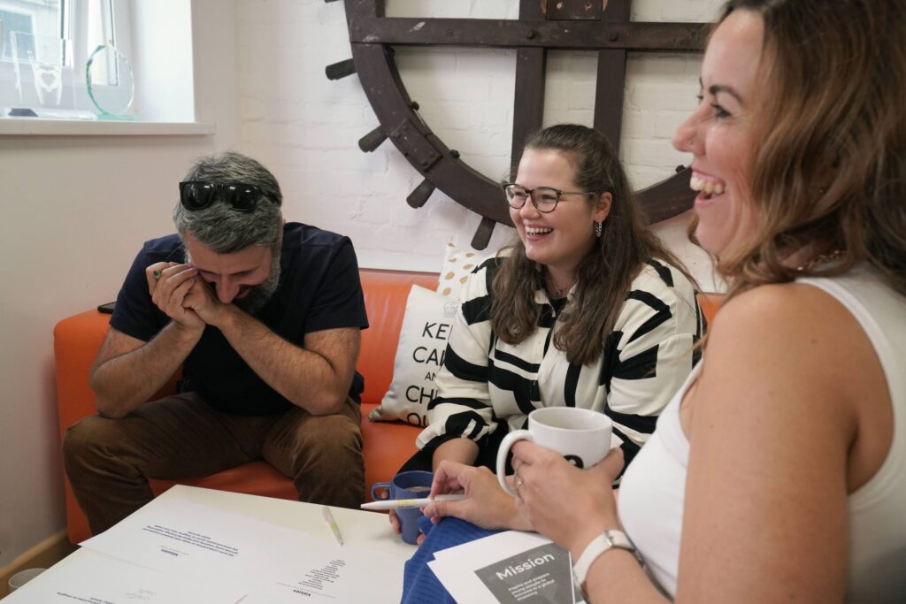 Three people sat on orange sofa chatting and working while laughing