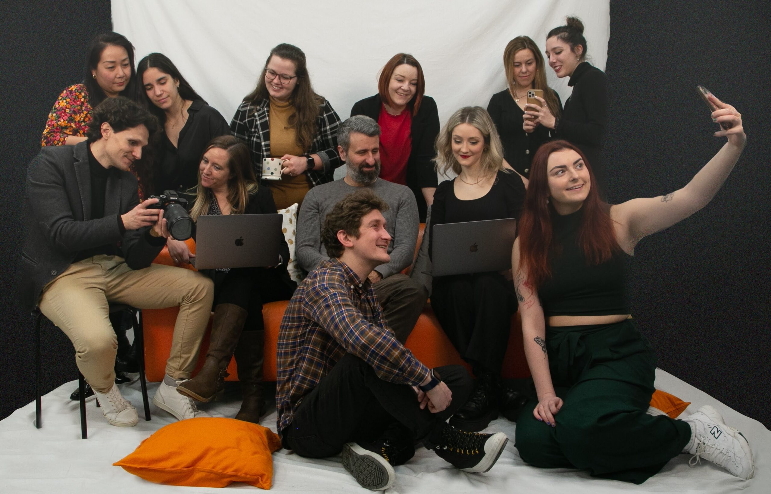 Group of people gathered on sofa, standing and sitting on floor. All looking at different devices together, smiling.