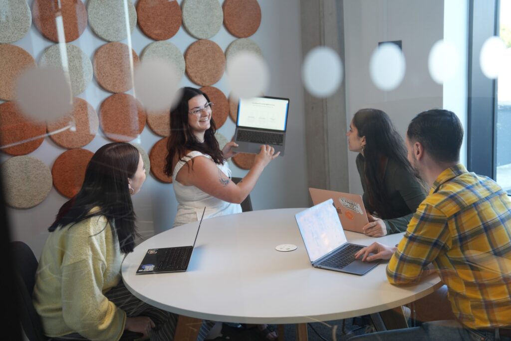Four people sat around a round table with laptops. One holds laptop up to show something on her screen to the others.